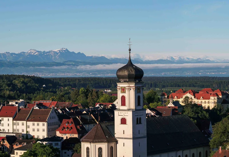 Kirche mit Bodensee Blick auf eine Kirche mit Zwiebelturm vor einer Stadtlandschaft und dahinter Bergen unter klarem Himmel.
